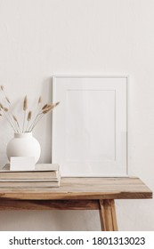 Portrait white picture frame mockup on vintage bench, table. Modern ceramic vase with dry Lagurus ovatus grass, books and busines card. White wall background. Scandinavian interior. Vertical.
