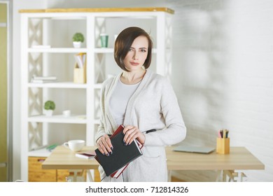 Portrait Of White Woman Holding Closed Book In Modern Office Interior. Knowledge And Information Concept 