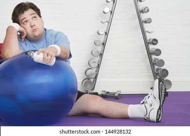 Portrait Of A Wistful Overweight Man Sitting On Floor With Exercise Ball In Health Club