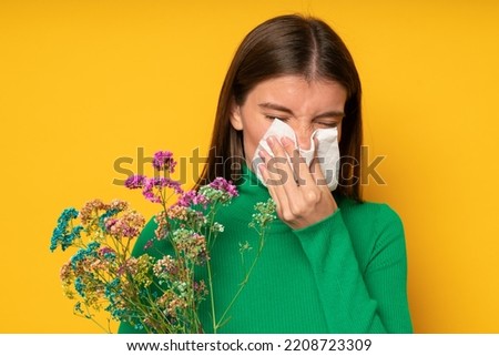 Portrait of woman allergic to wild spring flowers, holding bouquet in hands, blowing nose in handkerchief with closed eyes isolated on yellow studio background, dressed in green clothes