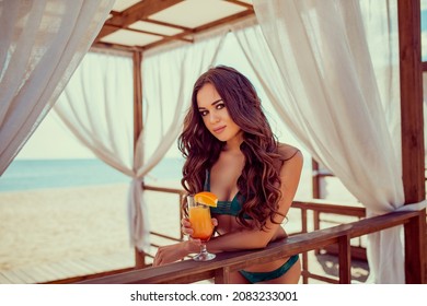 Portrait of woman in bikini with orange cocktail glass with straw and fruit posing at wooden bar of tent with white curtains. 