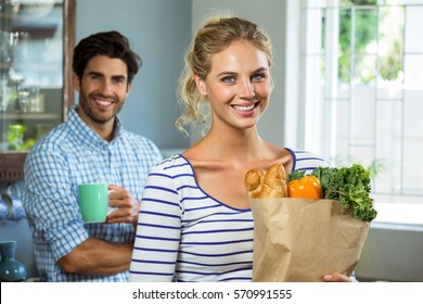 Portrait Of A Woman Carrying Grocery Bag While Man With Coffee Cup In The Kitchen At Home