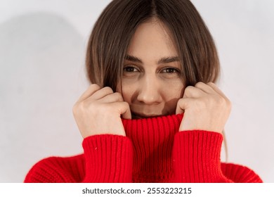 Portrait of a woman in close-up. A beautiful young woman covers her mouth with the collar of a red sweater holding it with her hands. - Powered by Shutterstock