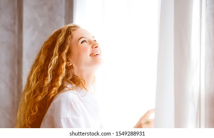 Portrait Of Woman Curly Blonde Hair Open Sheer Curtain And Looking Up Through The Window With Morning Light.Lifestyle Gorgeous Brunette Lady Happy Feeling With Soft Light While Peek Out The Window.