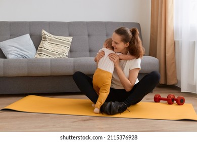 Portrait Of Woman With Dark Hair Wearing Black Leggins And White T Shirt Sitting On Yoga Mat And Hugging Her Baby Daughter Coming To Her While She Was Doing Sports At Home.