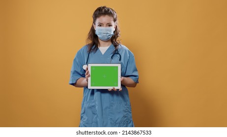 Portrait Of Woman Doctor Holding Tablet Computer With Green Screen Wearing Hospital Uniform And Covid Protection Mask In Studio. Medic Using Digital Touchscreen Device For Advertising Copy Space.