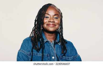 Portrait of a woman with dreadlocks looking at the camera with a smile while standing against a grey background. Stylish mature woman wearing a denim jacket and make-up in a studio. - Powered by Shutterstock