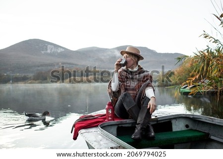 Portrait of woman dressed in poncho, sit in the boat, drink tea and look to the lake, enjoy life, happy in moment