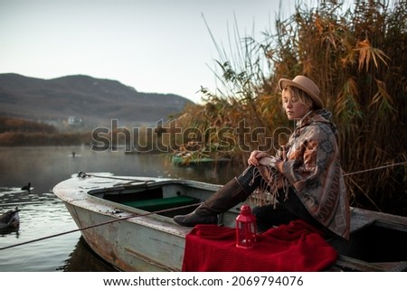 Portrait of woman dressed in poncho, sit in the boat and look to the lake, enjoy life, happy in moment