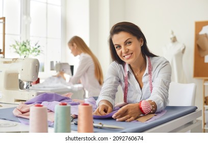 Portrait of woman dressmaker, fashion designer, tailor or seamstress at work in studio. Smiling woman looking at camera while working with fabric while sitting at her workplace in bright sewing studio