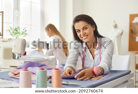 Portrait of woman dressmaker, fashion designer, tailor or seamstress at work in studio. Smiling woman looking at camera while working with fabric while sitting at her workplace in bright sewing studio