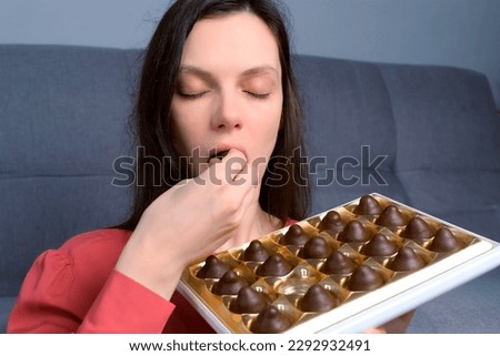 Portrait of woman is eating chocolate candies from box smelling them before it and smiling. Box of chocolates sweets set. Lots of truffles in a gift box. Bad habits, eating sweets.
