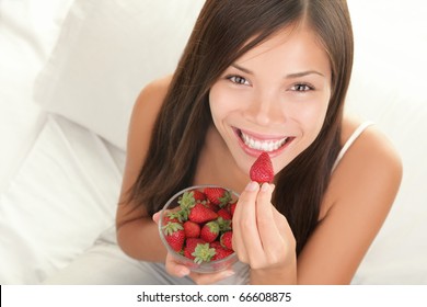 Portrait of woman eating strawberries. Healthy happy smiling woman eating strawberry inside in bed holding a bowl of strawberries. Gorgeous smile on mixed Caucasian Asian female model. - Powered by Shutterstock