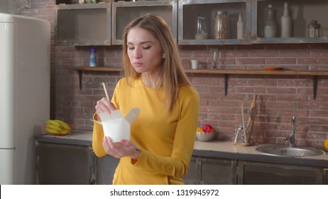 Portrait woman eating takeaway asian food from white box holding chopsticks. Happy adult lady relish take away chinese cuisine in home kitchen. Caucasian redhead lady has lunch or dinner alone in