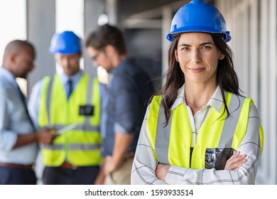 Portrait of woman engineer at building site looking at camera with copy space. Mature construction manager standing in yellow safety vest and blue hardhat with crossed arms. Successful architect.