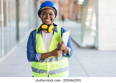 Portrait Of Woman Engineer At Building Site Looking At Camera With Copy Space. Mature Construction Manager Standing In Yellow Safety Vest And Blue Hardhat With Crossed Arms. 