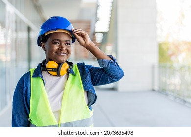 Portrait of woman engineer at building site looking at camera with copy space. Construction manager standing in yellow safety vest and blue hardhat. Successful confident architect at construction site