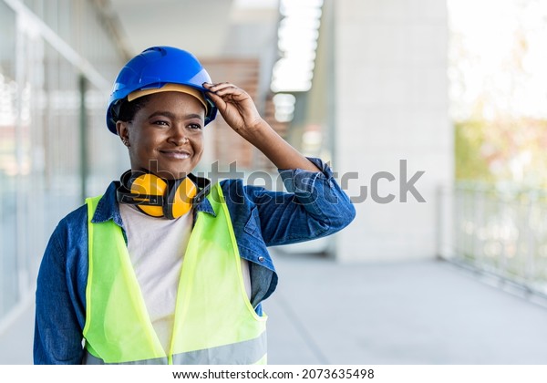 Portrait of woman engineer at building site looking\
at camera with copy space. Construction manager standing in yellow\
safety vest and blue hardhat. Successful confident architect at\
construction site