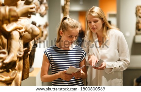 Portrait of woman with girl holding guidebook, standing in museum of ancient sculpture