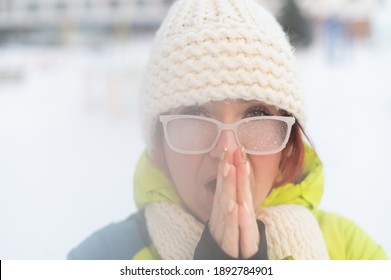 Portrait of a woman in glasses covered with hoarfrost. The girl is freezing and forgot gloves in very cold weather and blows on her bare hands - Powered by Shutterstock