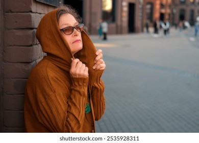 Portrait of a woman in glasses wearing a brown hooded sweater standing against a brick wall in an urban setting. Outdoor street photography. Woman 50 years old (fifty year old) - Powered by Shutterstock