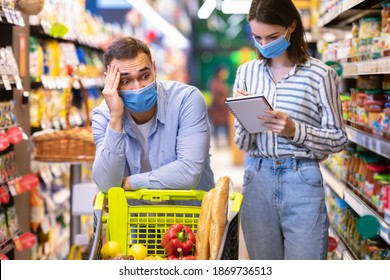 Portrait Of Woman Holding And Checking Grocery Shopping List, Writing In Notebook, Tired Bored Funny Man Leaning On Cart. Couple Buying Products, Standing In Supermarket, Wearing Medical Masks