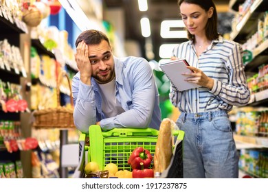 Portrait Of Woman Holding Checking Grocery Shopping List, Writing In Notebook, Tired Bored Funny Man Shocked By Price Leaning On Cart. Couple Buying Products In Supermarket With Trolley Full Of Food