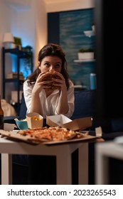 Portrait of woman holding delicious buger eating takeaway delivery food watching entertainment movie on television. takeout food, eating home, home delivery, burger delivery, beer bottle,