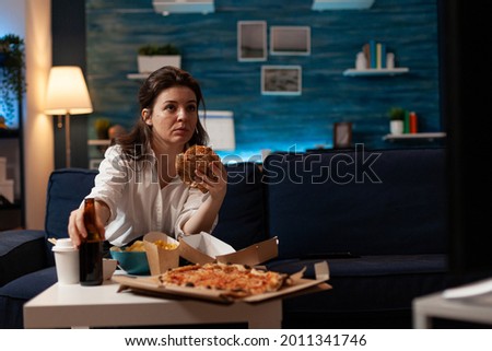 Portrait of woman holding delicious buger eating takeaway delivery food watching entertainment movie on television. Caucasian female sitting on couch tasting tasty junk-food enjoying night