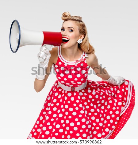 Portrait of woman holding megaphone, dressed in pin-up style red dress in polka dot and white gloves, on grey background. Caucasian blond model posing in retro fashion vintage studio shoot.