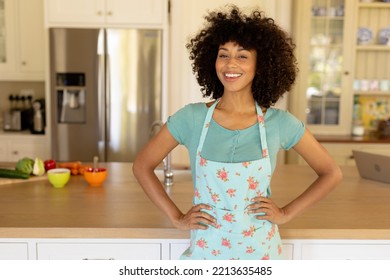 Portrait Of  Woman At Home, Standing In The Kitchen With Hands On Hips Wearing A Flowery Apron And Smiling To Camera