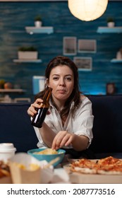 Portrait Of Woman Looking At Camera Holding Beer Bottle And Eating Potato Chips From Bowl At Table With Large Pizza And Takeaway Dinner. Office Worker On Couch Relaxing After Takeout Menu.