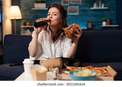 Portrait of woman looking into camera during fastfood lunch meal order relaxing on sofa late at night in living room. Caucasian female enjoying tasty burger, takeaway food home delivered