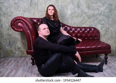 Portrait Of A Woman And A Man Seated On A Vintage Red Leather Sofa. Studio Portrait Of An Adult Couple In Retro Style