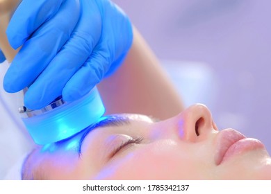 Portrait of woman on apparatus hardware phonophoresis face procedure with hyaluronic acid gel in cosmetology clinic, side view. Cosmetologist beautician doctor moving manipula with cooling blue light.