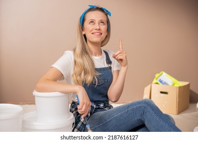 Portrait Of Woman On Brown Freshly Painted Wall, Advertising Acrylic Paint For Interiors And Ceilings. Girl Sits On Floor In Work Clothes And Points Upward With Finger To Spot On Inscription.