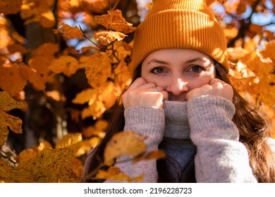 A portrait of a woman in an orange beanie in the orange leaves. The girl covers her face with a sweater because of the cold.