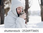 Portrait of a woman with a paper cup of hot drink in the winter forest. A young woman in a white fur coat and white hat enjoys a hot drink in the winter park. Copy space.