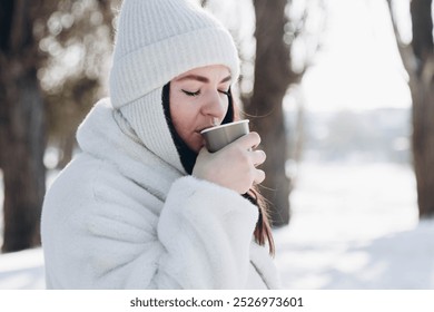 Portrait of a woman with a paper cup of hot drink in the winter forest. A young woman in a white fur coat and white hat enjoys a hot drink in the winter park. Copy space. - Powered by Shutterstock