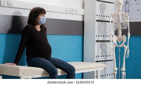 Portrait Of Woman With Pregnancy Belly Sitting On Bed In Office, Waiting To Attend Checkup Examination With Doctor. Pregnant Patient Expecting Child During Coronavirus Pandemic.