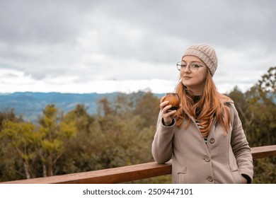 portrait of woman with red hair wearing glasses with winter clothes coat and beige hat drinking mate tea, gaucho drink yerba mate with forest and mountains background - Powered by Shutterstock