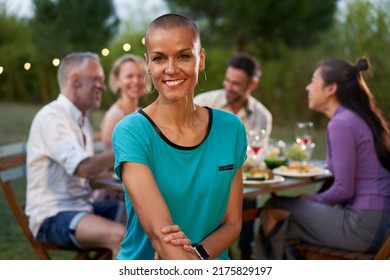 Portrait Of A Woman With Shaved Head Looking At The Camera. Group Of Friends Dining Outside On A Terrace Restaurant Or Backyard Home. Friends Having Fun Eating And Drinking On A Summer Night.