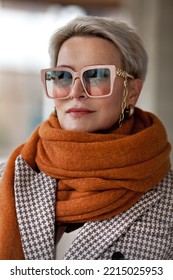 Portrait Woman Short Blonde Hair In Warm Knitted Ochre Scarf, Double-breasted Jacket And Fashionable Glasses. Fashion Style Autumn Or Spring, Female Fashionista 50 Years Old