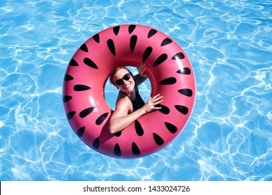 Portrait Of Woman With Short Hair Swimming In A Pool, Look Laughing Through Pink Floatie Inflatable Doughnut, Blue Water. Funny Mood.                              