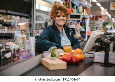 Portrait Of A Woman Sitting Behind Checkout Counter Smiling At Camera. Supermarket Cashier At Checkout.