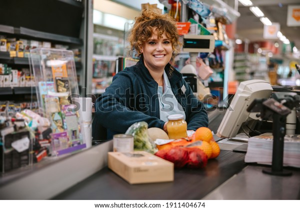 Portrait of a woman\
sitting behind checkout counter smiling at camera. Supermarket\
cashier at checkout.