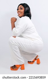 Portrait Of A Woman Squatting. Smiling African Lady Wearing All White Dress And Orange Heels. 