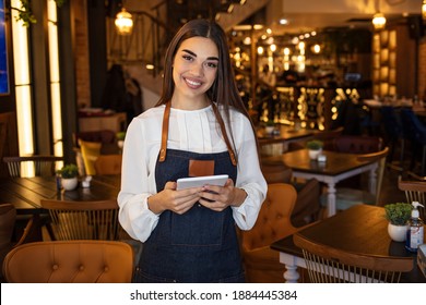 Portrait of woman standing at doorway of her store holding digital tablet. Cheerful waitress waiting for clients at coffee shop. Successful small business owner in casual clothing looking at camera.
