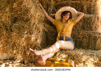 Portrait of a woman with a straw hat, yellow shirt, jean shorts and a yellow suitcase sitting in a hayloft.