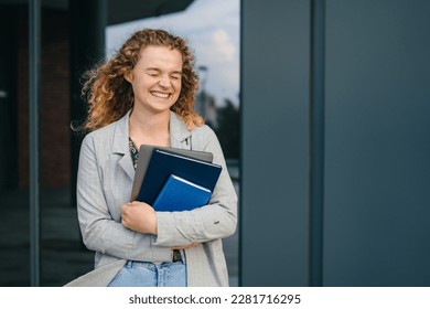 Portrait of woman student standing outdside the university campus wearing jacket, holding books and laptop having coffee break after lecture. Enjoying college